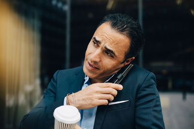 Mature businessman talking from smart phone while holding disposable coffee cup against building in city