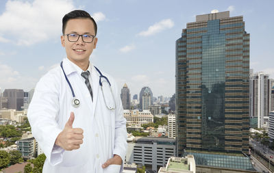 Portrait of confident doctor gesturing thumbs up against buildings in city