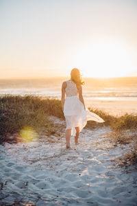 Rear view of young woman walking at beach against clear sky during sunset