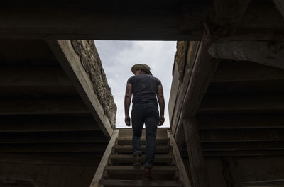 Rear view of adult man in cowboy hat climbing up wooden stairs, indoor toward rooftop