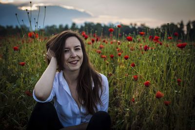 Portrait of smiling young woman sitting by poppy flowers on field