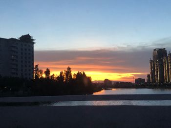 Silhouette buildings by river against sky during sunset