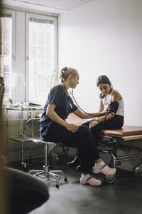 Female nurse examining blood pressure of girl sitting on bed in clinic