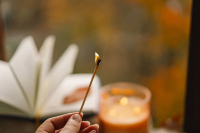 Hand with burning match lighting a candle on the windowsill with autumn decor