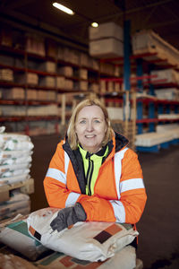 Portrait of happy blue-collar worker in protective workwear leaning on package in distribution warehouse