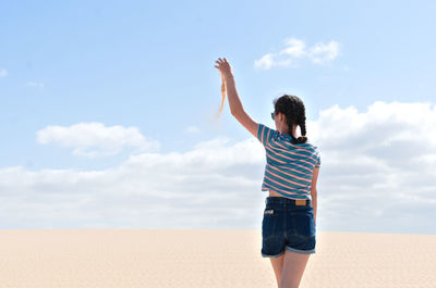 Rear view of teenage girl holding sand while standing on beach against sky