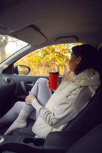 Traveler woman in a white vest is sitting in a car with a red paper cup of coffee in the autumn