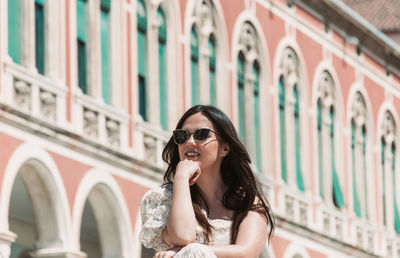 Portrait of beautiful young woman with long dark hair, sitting in square with beautiful architecture