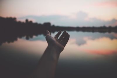 Cropped hand over lake against sky during sunset