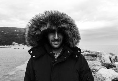 Portrait of smiling young man in warm clothing standing at beach against cloudy sky