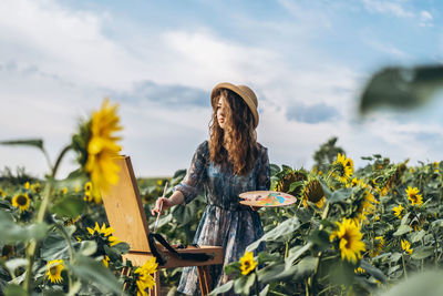 A young woman with curly hair and wearing a hat is painting in nature. 
