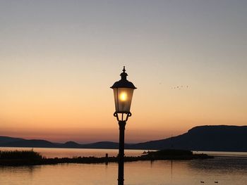 Silhouette street light by lake against sky during sunset