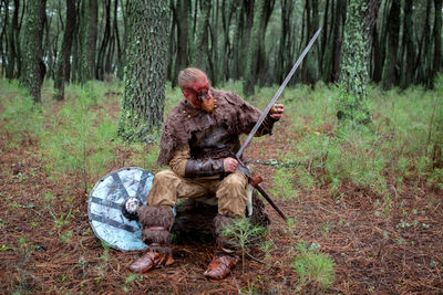 Full length of warrior holding sword sitting at forest