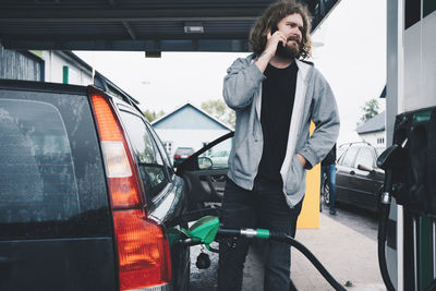 Young man talking on mobile phone while refueling car at gas station