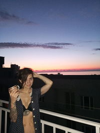 Woman standing by building against sky during sunset