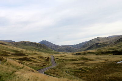 Road amidst grassy field against cloudy sky at cairngorms national park