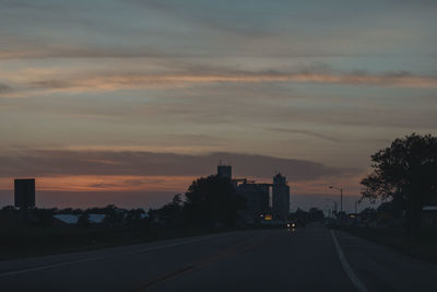Road by silhouette trees against sky at sunset