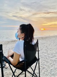 Rear view of woman sitting on chair at beach against sky during sunset