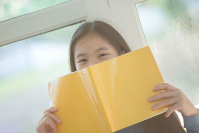 Portrait of girl with face covered by book against window at home