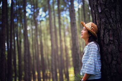 Side view of young woman looking away while standing by tree trunk in forest