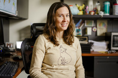 A woman professor sits at her office desk smiling with direct gaze