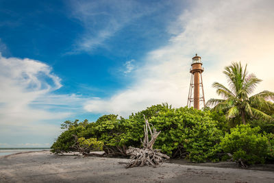 Low angle view of lighthouse amidst trees by beach against cloudy sky