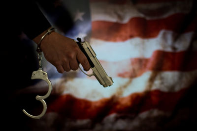 Cropped hand of male criminal holding gun against american flag