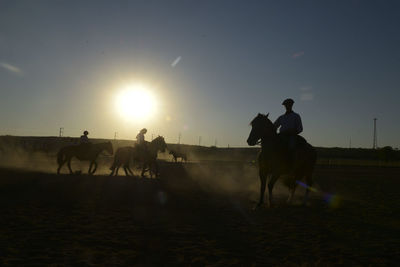 People riding horses on field against sky during sunset