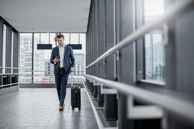 Young businessman in a passageway with cell phone, earbuds and rolling suitcase on the go