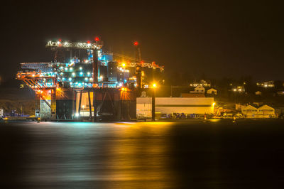 Illuminated factory by sea against sky at night