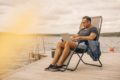 Full length of mature man using laptop while sitting on deck chair at jetty