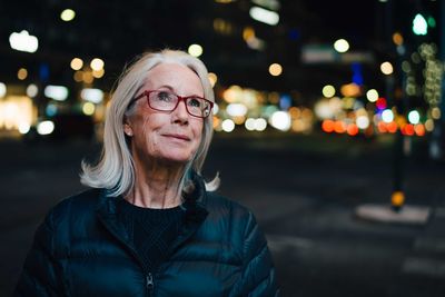 Wrinkled smiling woman looking up while standing in illuminated city at night