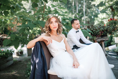 Portrait of beautiful bride sitting on park bench with bridegroom in background