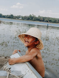 Smiling shirtless boy holding boat in river against sky