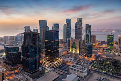 Modern buildings in city by river against sky during sunset