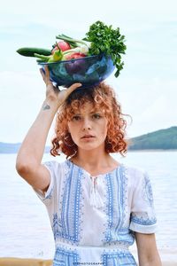 Portrait of young woman carrying bowl with vegetables on head against sea