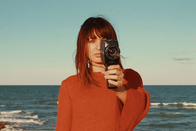 Portrait of woman holding camera at beach against sky