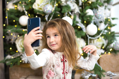 Portrait of girl holding mobile phone at christmas tree