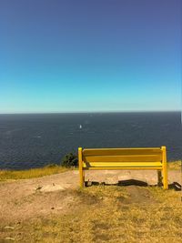 Empty bench by sea against clear blue sky