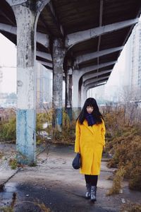 Full length of woman wearing yellow overcoat while walking under bridge in city