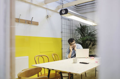 Young man using smart phone and laptop while sitting at desk in board room