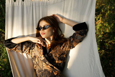 Portrait of young woman wearing sunglasses standing against curtain