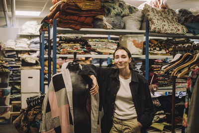 Portrait of female fashion designer with hand in pocket leaning on mannequin at workshop