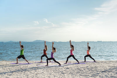 Young friends practicing yoga in warrior position on shore at beach during sunny day