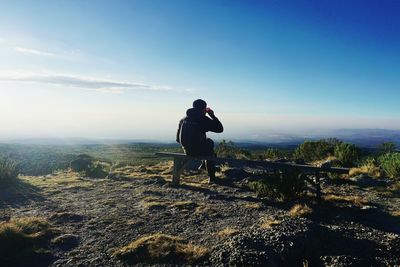 Rear view of man having drink while sitting on bench at mountain against sky