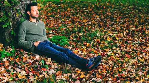 Young man looking away while sitting against tree trunk during autumn