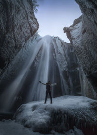 Rear view of man with arms outstretched standing against waterfall