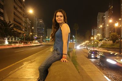 Portrait of young woman sitting at roadside in illuminated city at night