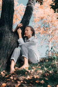 Woman sitting on grass by tree trunk during autumn