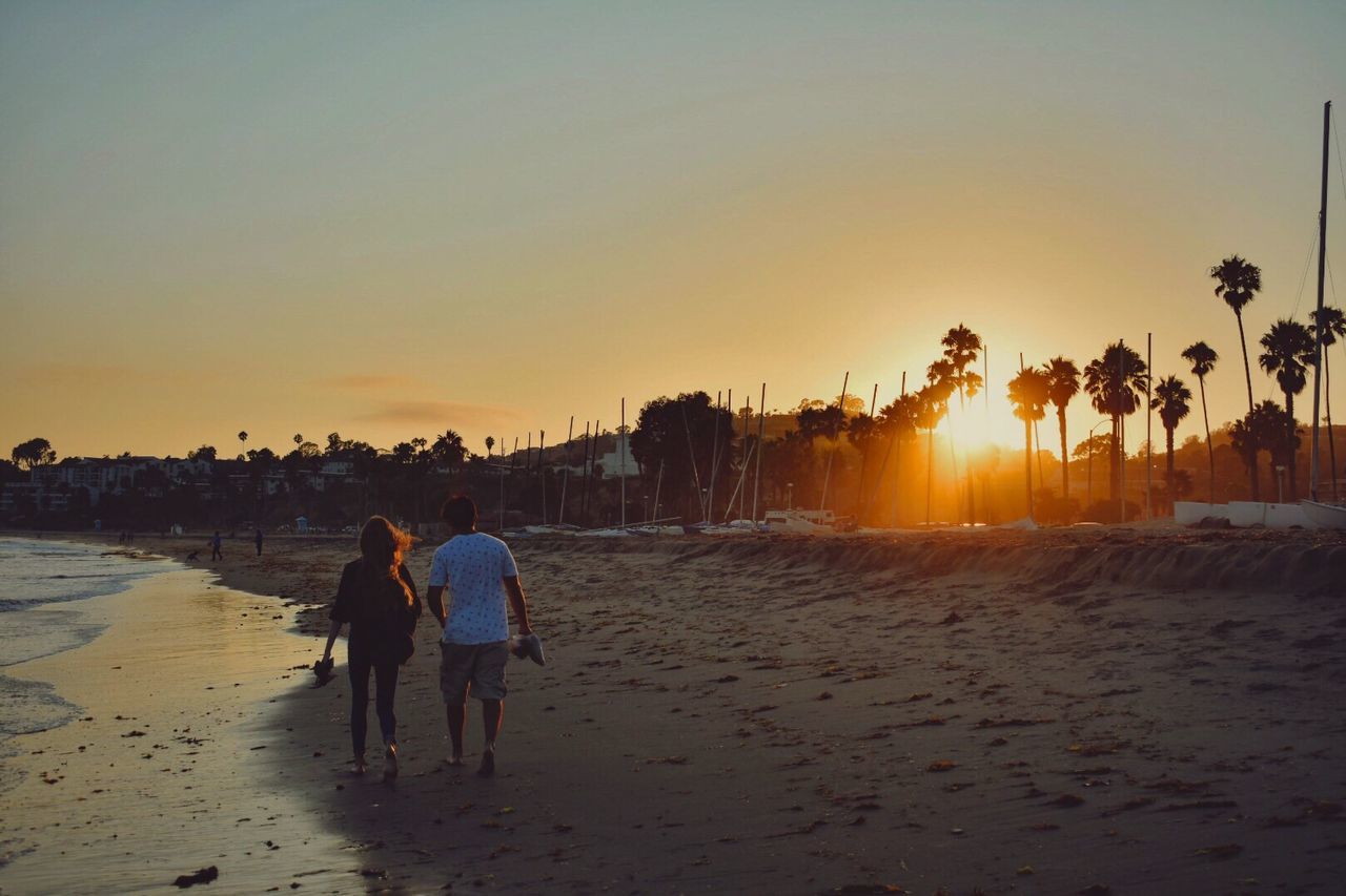 sunset, beach, real people, sand, togetherness, walking, full length, leisure activity, men, rear view, lifestyles, women, vacations, sun, two people, nature, sea, sunlight, outdoors, sky, weekend activities, beauty in nature, standing, bonding, clear sky, scenics, tree, day, adult, people